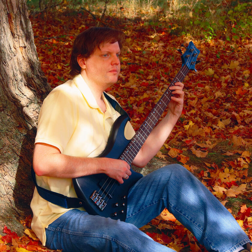 I am sitting beneath an oak tree in autumn in a bed of red, orange and yellow leaves. I am wearing a yellow polo shirt and jeans with a piccolo bass guitar in my lap, in playing position.