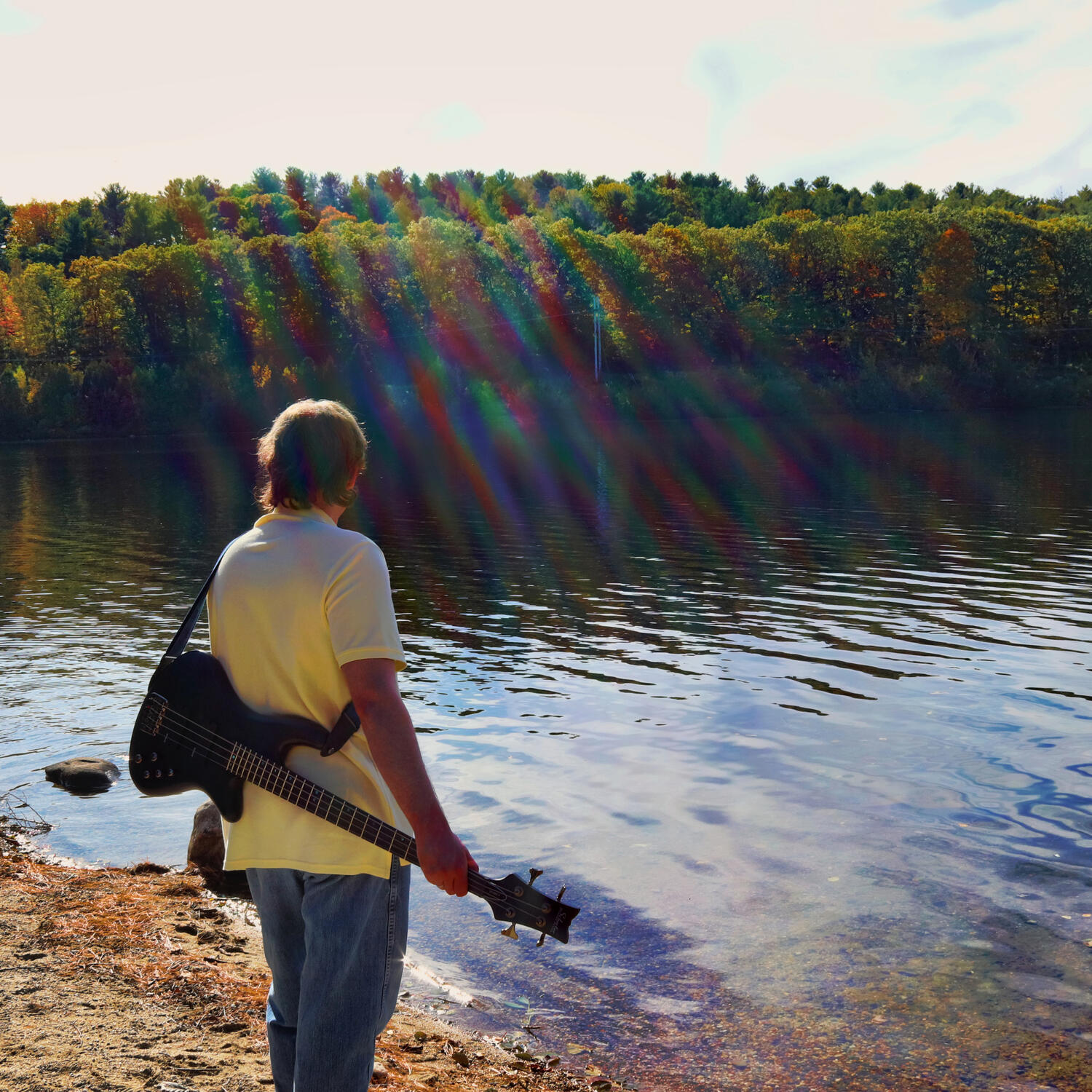 I am standing with my back to the camera and looking towards a body of water while wearing a yellow polo shirt, jeans and have a black bass guitar turned over my back so that the strings are facing the camera. The shore is mostly red sand and rocks while a