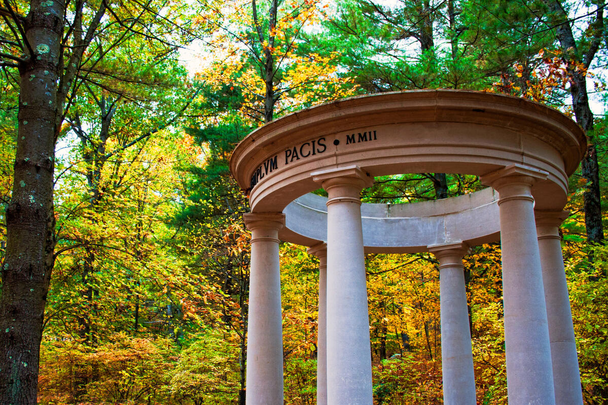 A Greco-Roman styled circular temple with doric pillars sits beneath an enclave of pine trees. Deciduous trees, green with gold colours coming in sit around the structure in the background. A pile of red, orange, and brown leaves lie on the forest floor in
