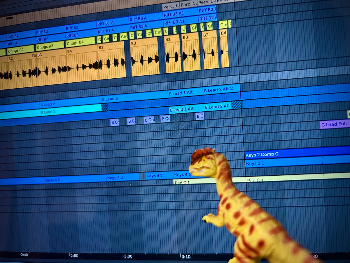 A green Dilophosaurus with brown spots stands before a computer screen with Ableton Live open. They are looking over various audio and MIDI tracks labelled in yellow, orange, and various shades of blue as they mix the pictured recording.