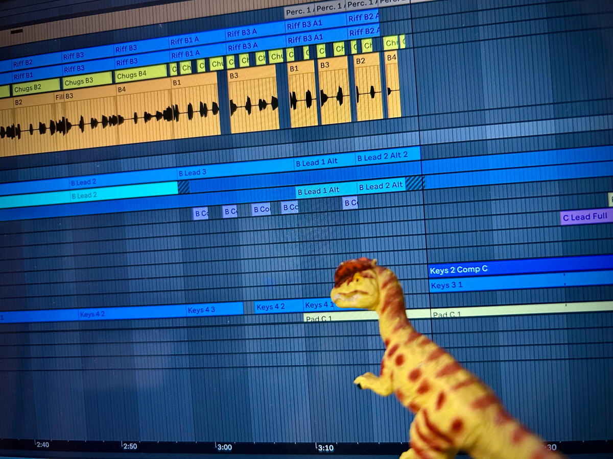 A green Dilophosaurus with brown spots stands before a computer screen with Ableton Live open. They are looking over various audio and MIDI tracks labelled in yellow, orange, and various shades of blue as they mix the pictured recording.