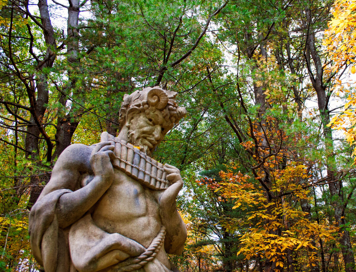 A statue of Pan with pipes in hand against an autumn forest of deciduous and fir trees.