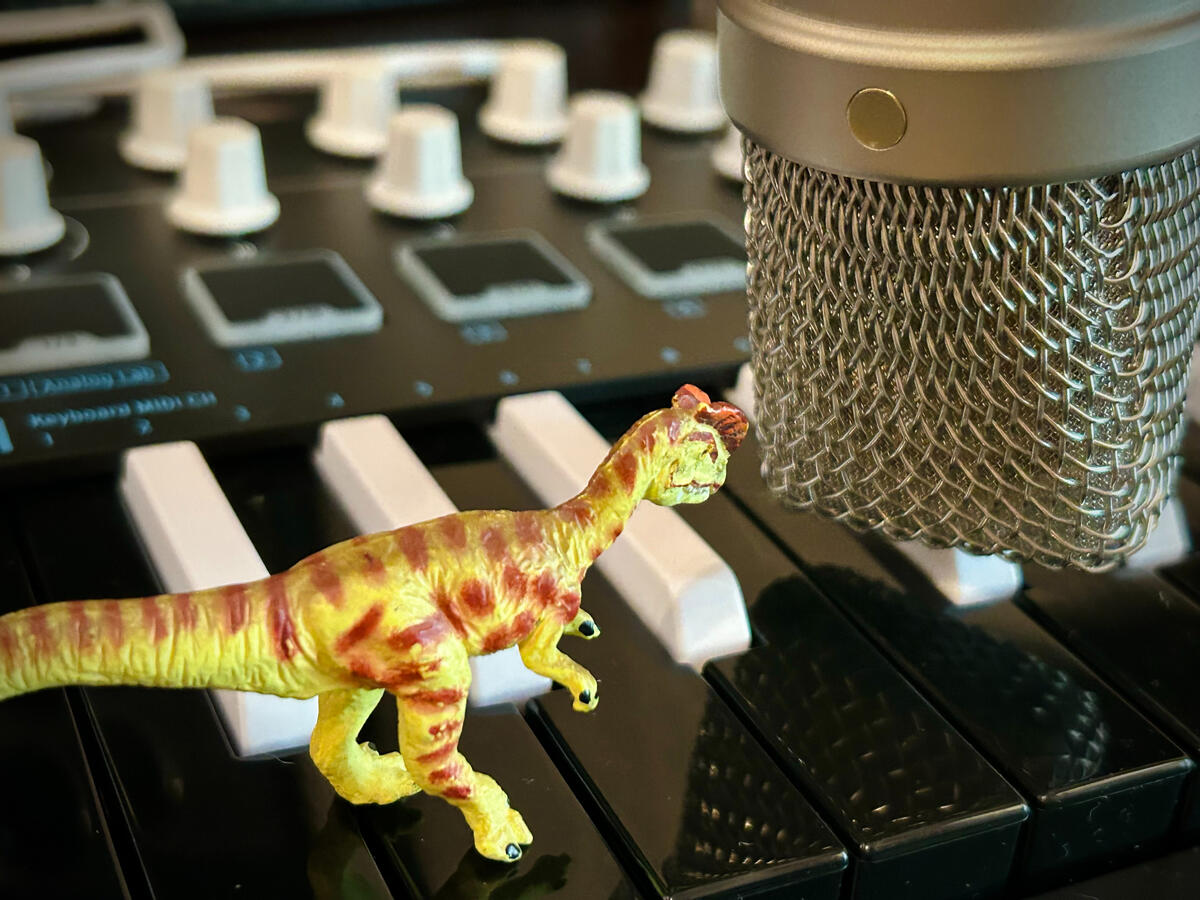 A green Dilophosaurus with brown spots stands on a mini piano keyboard with inverted colours and speaks into a golden Rhode condenser microphone with some hot takes. The controller's white knobs and pads are visible in the background.
